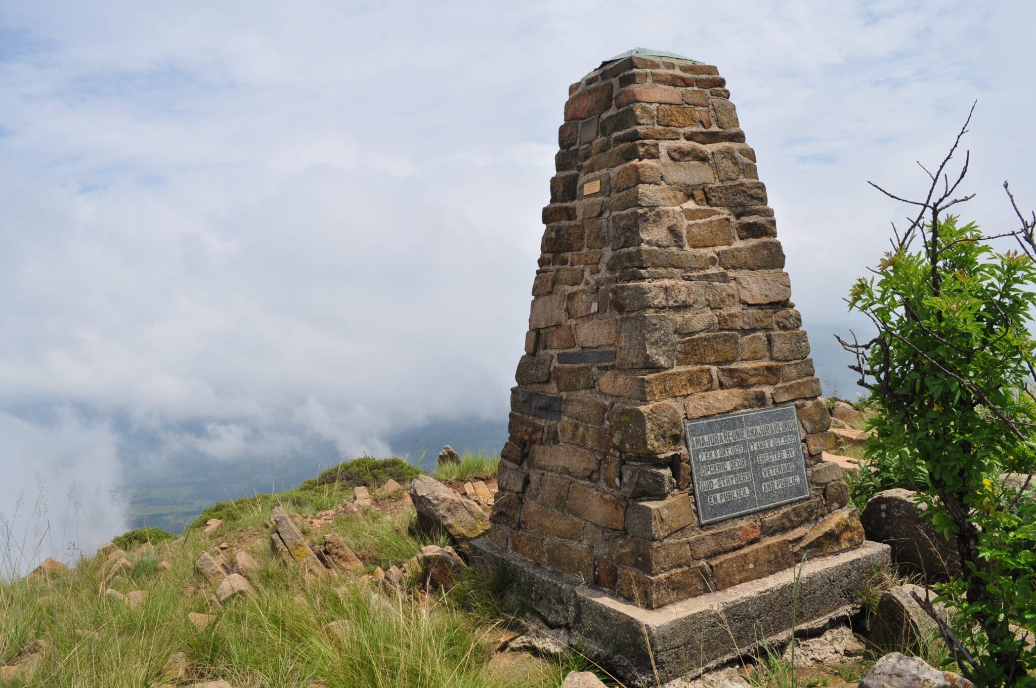 A stone monument with a plaque stands on a grassy hilltop surrounded by rocks