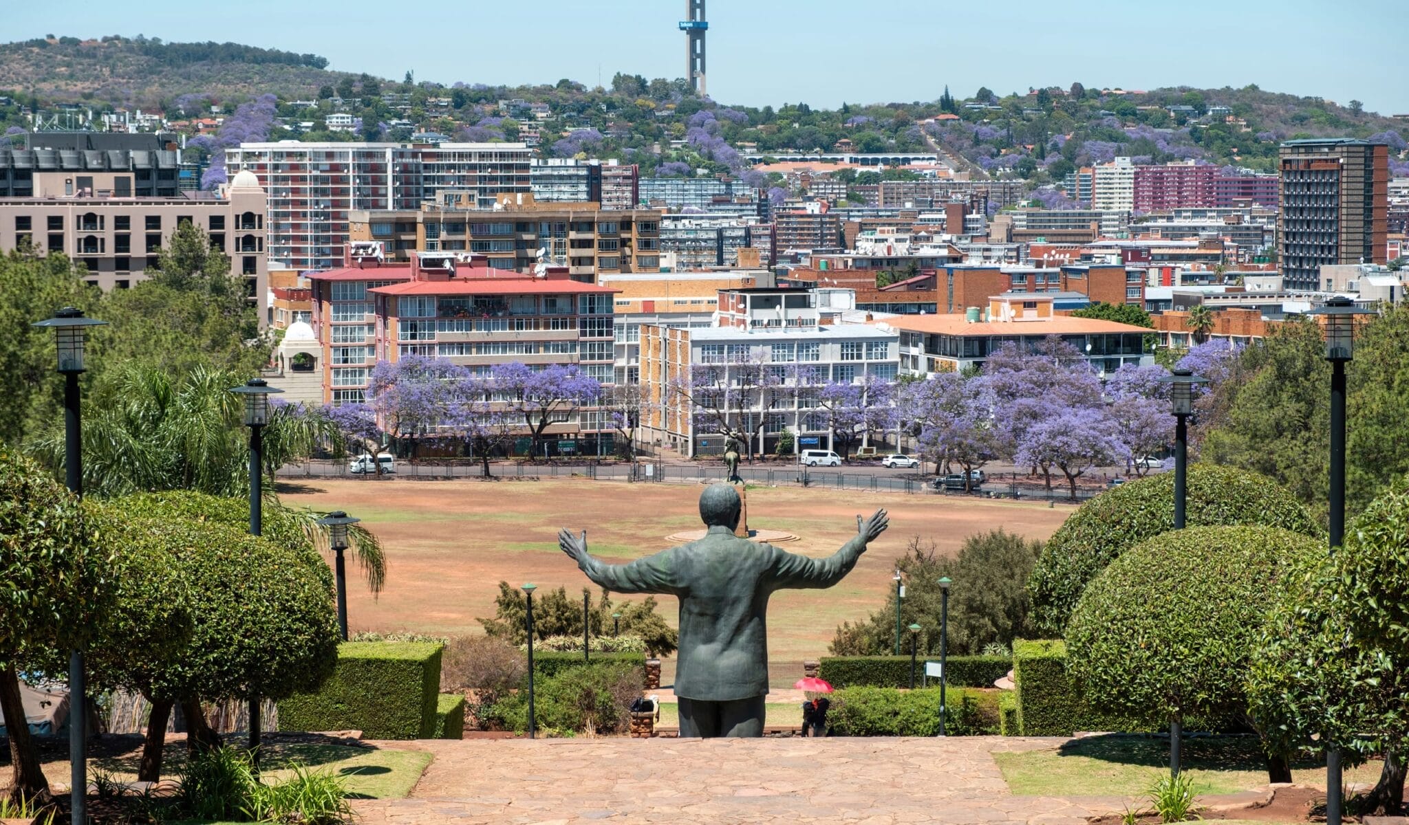 nelson-mandela-statue-rear-view-and-the-union-buil-2026-01-06-10-10-25-utc A statue with outstretched arms faces a cityscape filled with buildings and blooming jacaranda trees