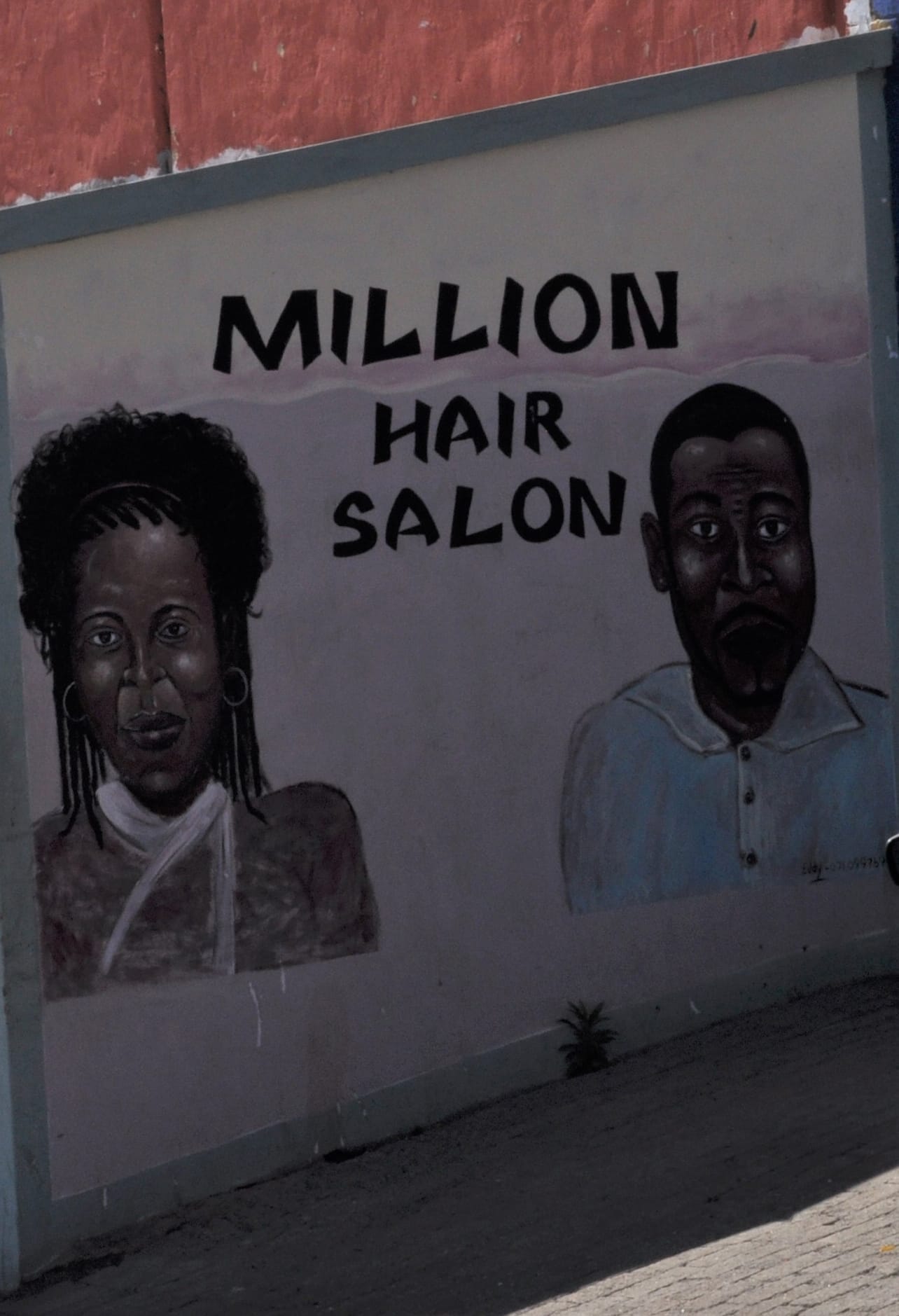 A mural on a wall reads “MILLION HAIR SALON” with painted portraits of a woman with curly hair on the left and a man with short hair on the right. The wall is light-coloured with a red upper section and pavement below.