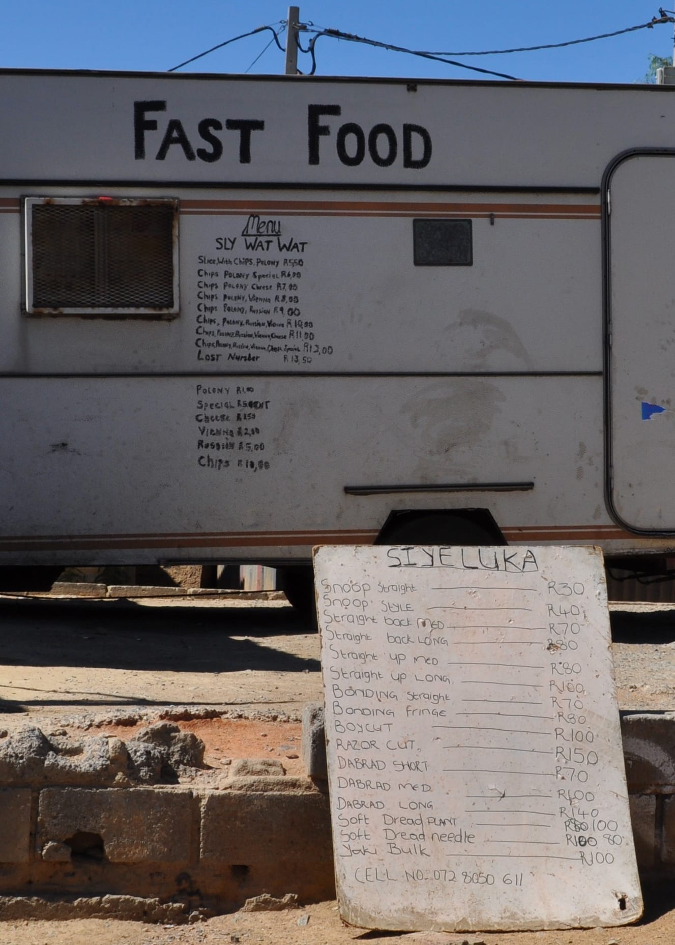 A food van with handwritten menus on its side and a whiteboard on the ground lists various fast food items with prices, including wors, steak, and boerewors rolls. The van is parked outdoors on a dirt surface under a clear blue sky.