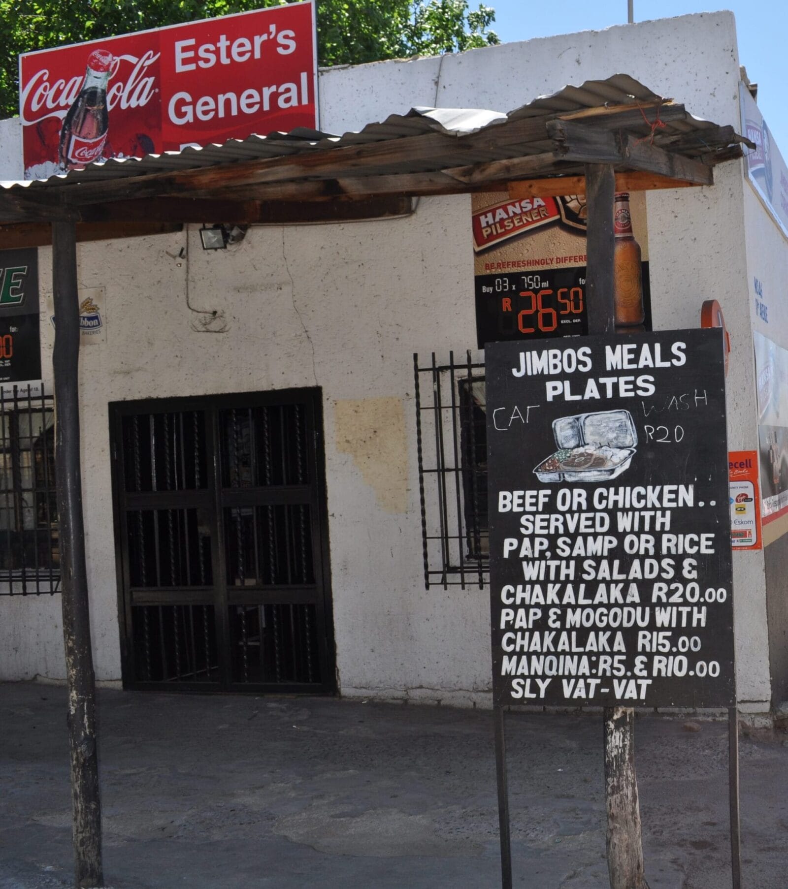 A small, white building has a Coca-Cola sign reading “Ester’s General.” Outside, a chalkboard advertises “Jimbos Meals Plates,” offering beef or chicken with sides and prices listed. There’s a barred entrance and a rusted metal roof above the sign.