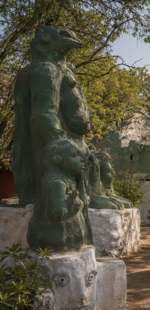 A tall, green, abstract sculpture with animal-like and human features stands outdoors among trees, accompanied by smaller busts on white, painted plinths, one of which has a painted face. A hut with a thatched roof is visible in the background.