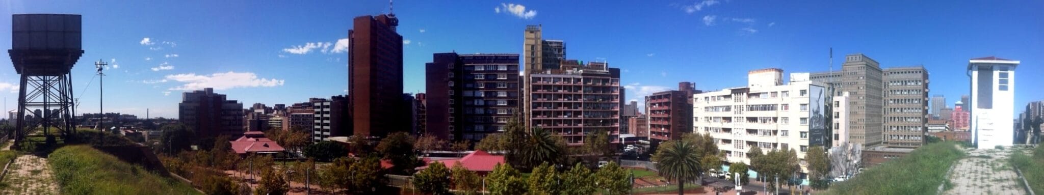 A panoramic cityscape under a clear blue sky, featuring tall modern and older buildings, trees