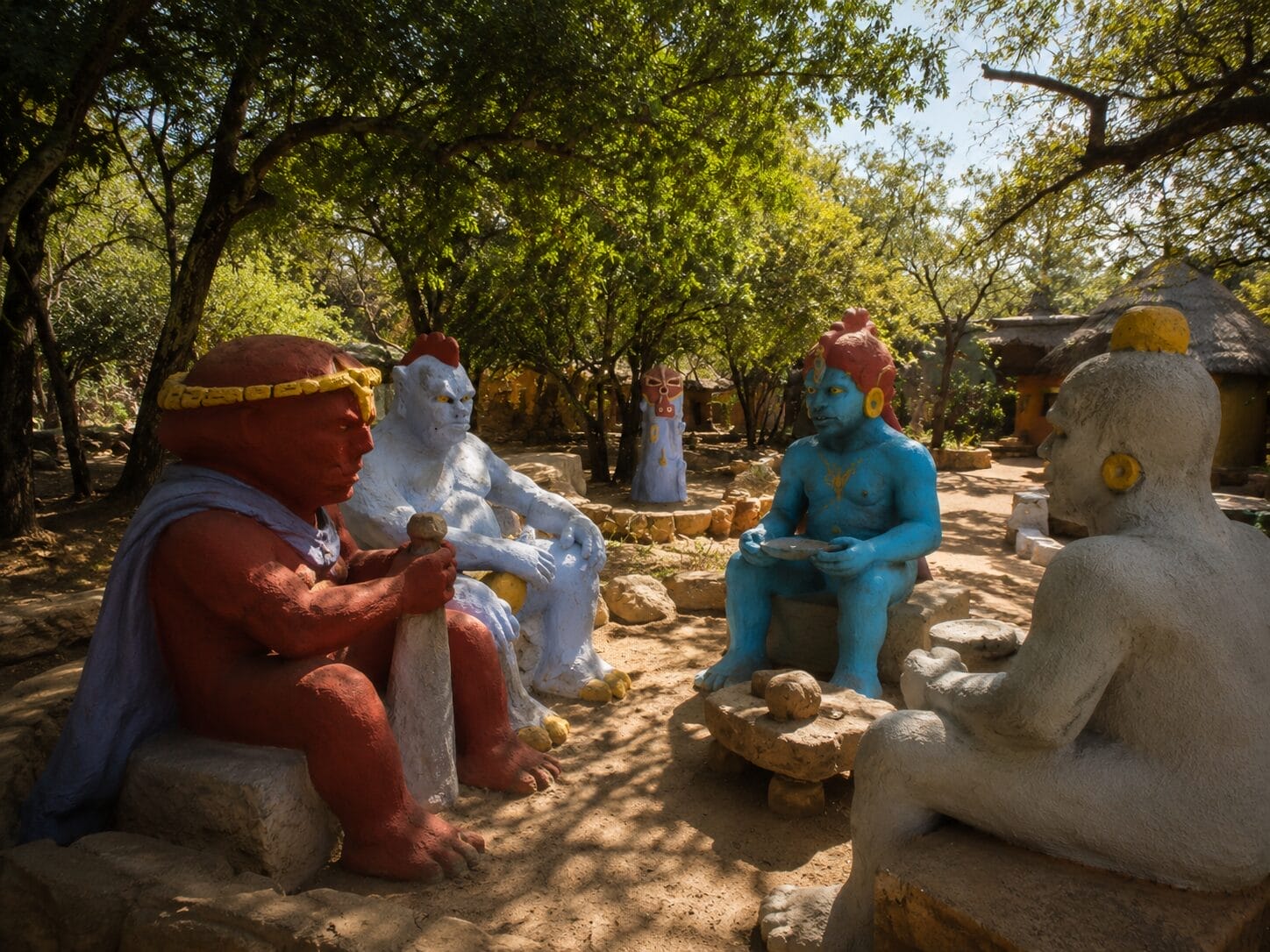 Four colourful life-sized statues of human figures sit in a circle outdoors, surrounded by trees
